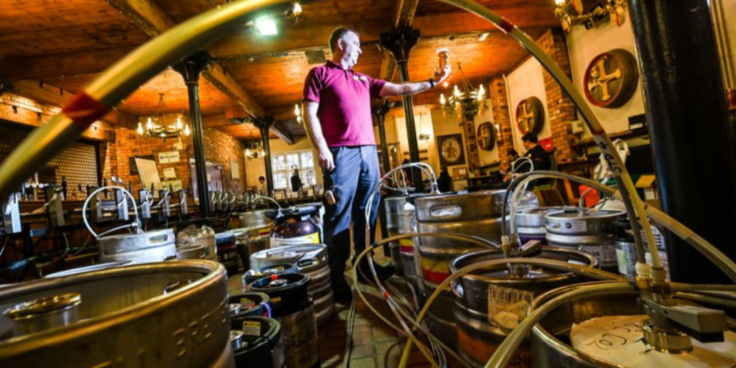 Man surrounded by brewing equipment who is holding a glass of beer
